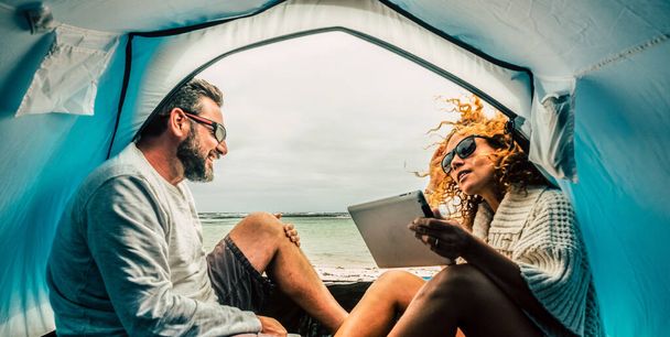 Couple adulte caucasien sourire et profiter des loisirs à l'intérieur d'une tente dans le camping gratuit à la plage avec vue sur la mer. Concept de vacances alternatives touristiques et de voyage. Femme utilisant la tablette et la connexion - Photo, image