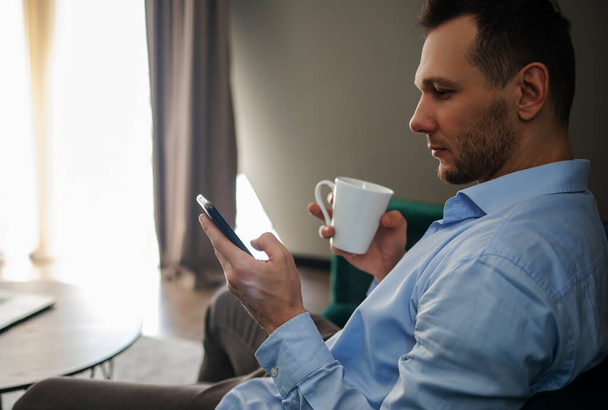 счастливый мужчина со смартфоном сидит на диване и держит чашку чая. - Фото, изображение