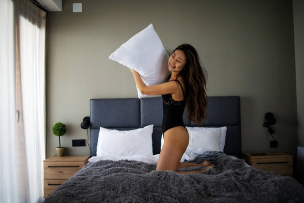 Счастливая молодая красивая азиатка прыгает на кровати дома, обнимая подушку. - Фото, изображение