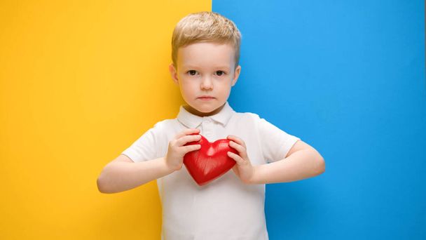 Child is pacifist. Call to love, hands shape of heart, Ukraine with love. Portrait little blond boy expresses love, presses red heart with his hands to his chest, standing on blue-yellow background. - Foto, Bild