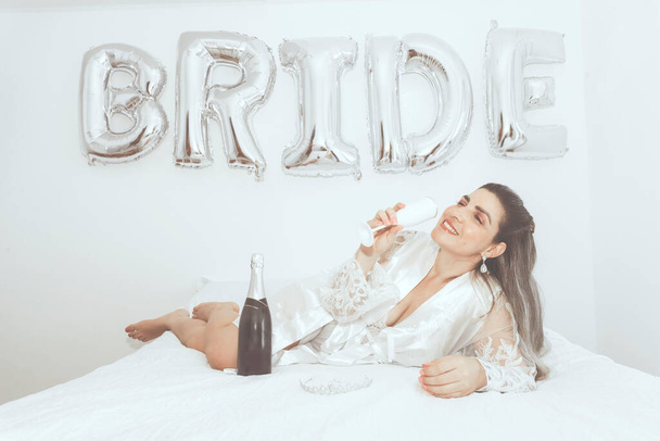 Woman lying in bed with glass of wine in hand and bottle of drink against white background written Bride. Salvador, Bahia, Brazil. - Фото, изображение