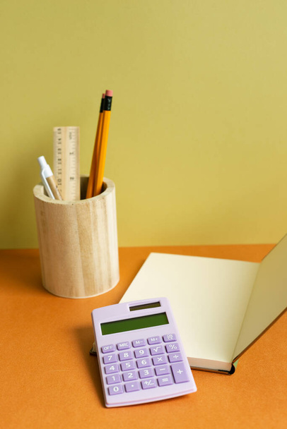 Cuaderno, calculadora, portalápices en escritorio naranja. fondo amarillo de la pared. espacio de trabajo - Foto, Imagen
