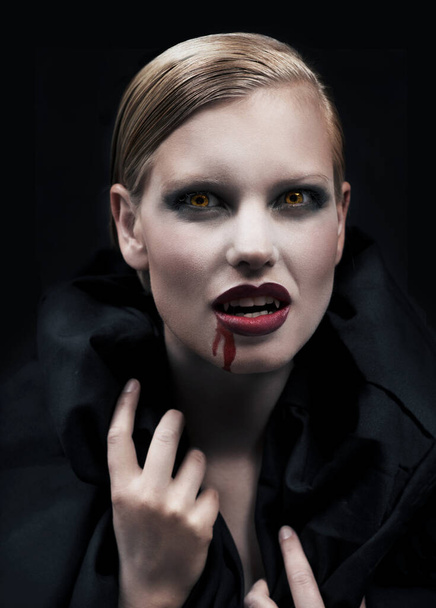 Fueled by a desire to feed. A provocative female vampire with blood running down her chin against a dark background. - Foto, Bild