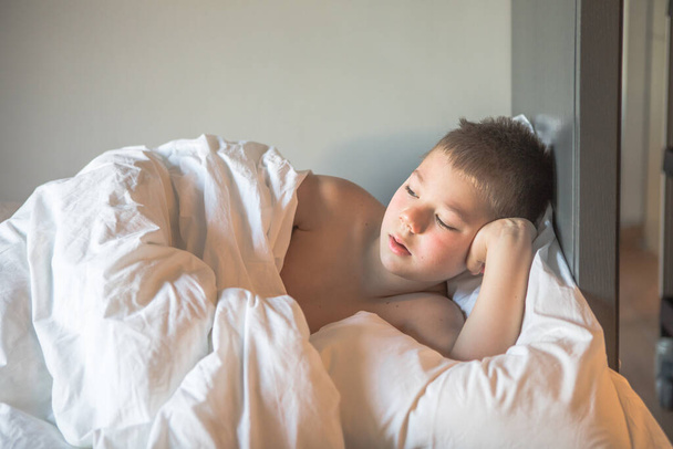 7歳の子供はベッドに横になり、眠い子供は朝起きてベッドルームで朝の光で、悲しい少年は部屋のベッドの上に横たわっています - 写真・画像