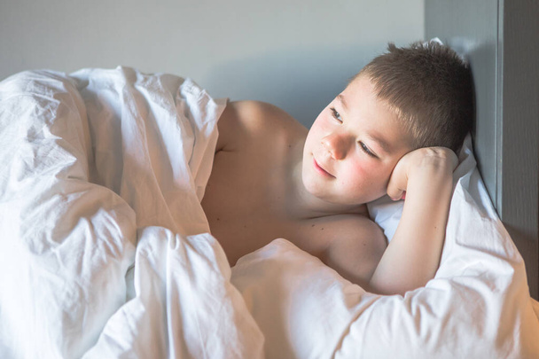 Kid 7 anos deitado na cama, criança sonolenta acordando pela manhã em seu quarto de cama com luz da manhã, menino triste jaz em uma cama no quarto - Foto, Imagem