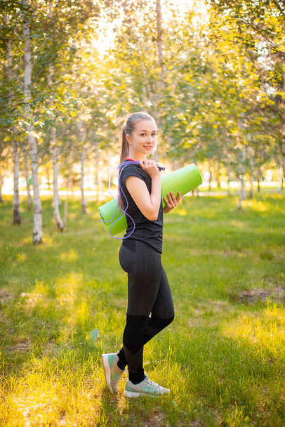 Portrait of a beautiful smiling young woman in a sports uniform with a jump rope and a yoga mat in her hands, posing against the backdrop of a park. - Zdjęcie, obraz