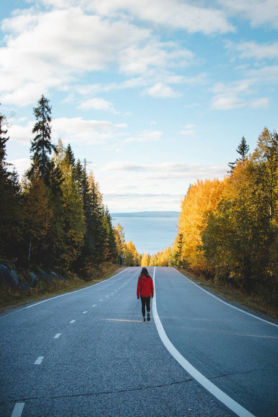 voyageuse brune active dans une veste rouge trouve son propre chemin dans la vie. Un explorateur de 20 ans se promène le long de la route principale à travers les bois d'automne. Région de Kainuu, Finlande. - Photo, image