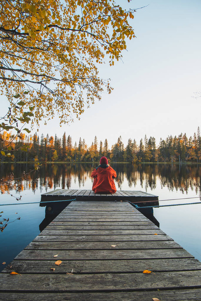 Pensive girl w czerwonej kurtce siedzi na końcu drewnianego molo z widokiem na spokojną wodę, jak słońce w Sotkamo, region kainuu, Finlandia. Krajobraz skandynawski. - Zdjęcie, obraz