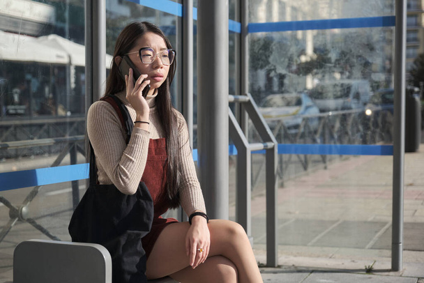 Młoda Azjatka rozmawia przez telefon czekając na przystanek autobusowy. - Zdjęcie, obraz