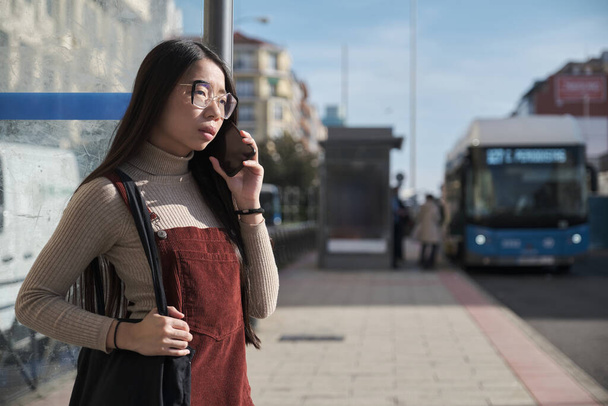 Young asian woman talking on her phone while waiting for the bus at bus stop. - Foto, afbeelding