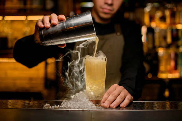 hand of bartender holds shaker and gently pours a steaming cocktail into a glass with ice pieces on the bar counter - Fotó, kép
