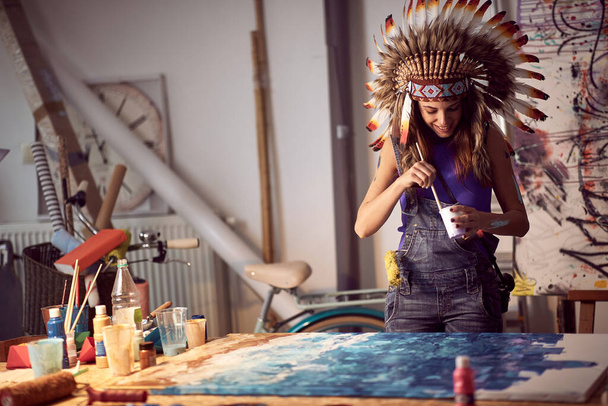 A young female artist with a war bonnet in a pleasant atmosphere in the studio is mixing and preparing the paint for a new painting. Art, painting, studio - 写真・画像