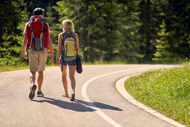 Couple walking on road. Man and woman with backpacks in forest on trail. Hiking, lifestyle, nature concept - 写真・画像