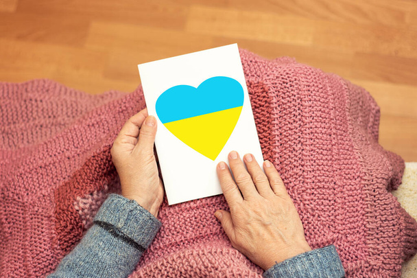 Old woman grandmother holding a card in her hands with a heart symbol in the color of the flag of Ukraine. We pray for Ukraine. - Foto, afbeelding