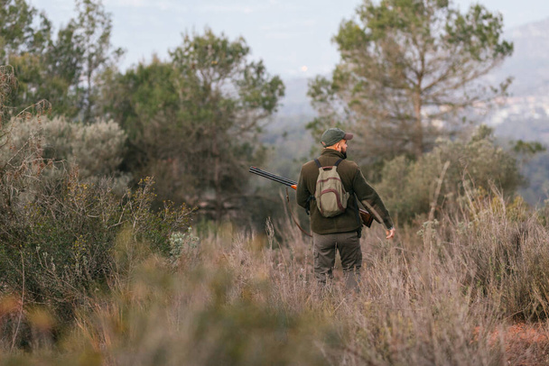 Αρσενικό κυνηγός κρατώντας ένα όπλο ενώ στέκεται στο πεδίο σε εξωτερικούς χώρους ψάχνει για ένα θήραμα. - Φωτογραφία, εικόνα