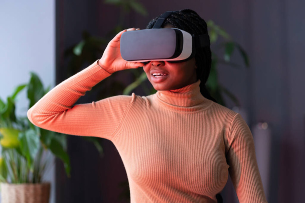 Mujer africana sonriente en casco VR sentada en interiores con luz de neón roja, viajando en 360 grados - Foto, Imagen