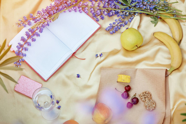 Otwórz notatnik makieta z kieliszkiem wina, łubin fioletowe kwiaty, jabłko, banany, wiśnia, halva, ciasteczka na złotym tle. Letni piknik. Miejsce na tekst. Bliskość natury, koncepcja samopoznania - Zdjęcie, obraz