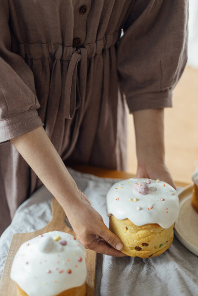 Женщина держит испеченный стильный пасхальный торт с сахарной глазурью и брызгами. Домашний пасхальный хлеб в руках на фоне деревенского стола в номере. Счастливая Пасха! - Фото, изображение