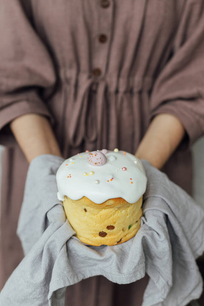 Домашний пасхальный хлеб в руках на фоне деревенской комнаты. Счастливой Пасхи! Женщина, держащая свежеиспеченный стильный пасхальный торт с сахарной глазурью и брызгами в льняной салфетке - Фото, изображение