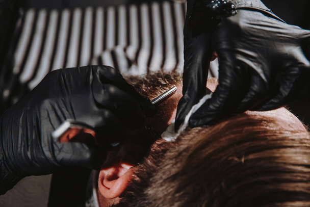 Bearded man getting haircut by hairdresser with dangerous razor at barbershop - 写真・画像