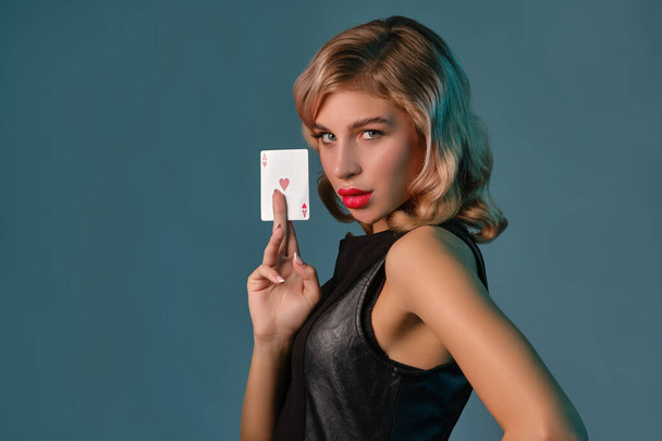 Blonde girl in black leather dress showing ace of hearts, posing against blue background. Gambling entertainment, poker, casino. Close-up. - Foto, immagini