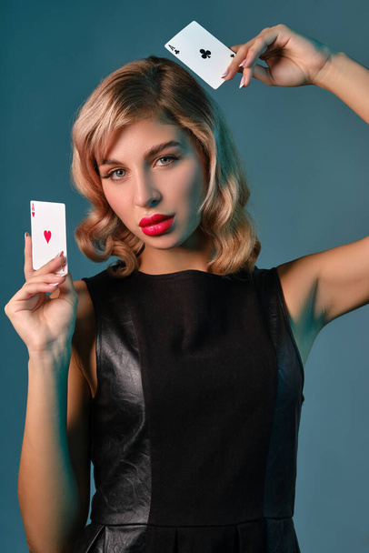 Blondes Mädchen im schwarzen Lederkleid, das zwei Spielkarten zeigt und vor blauem Hintergrund posiert. Glücksspiel, Poker, Casino. Nahaufnahme. - Foto, Bild