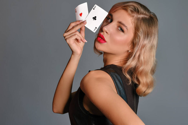 Blonde girl in black dress showing two playing cards, posing sideways against gray background. Gambling entertainment, poker, casino. Close-up. - Fotó, kép