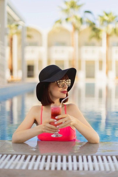Dáma v růžových plavkách v resortním bazénu, koupání a vychutnávání osvěžujícího studeného koktejlového nápoje - Fotografie, Obrázek