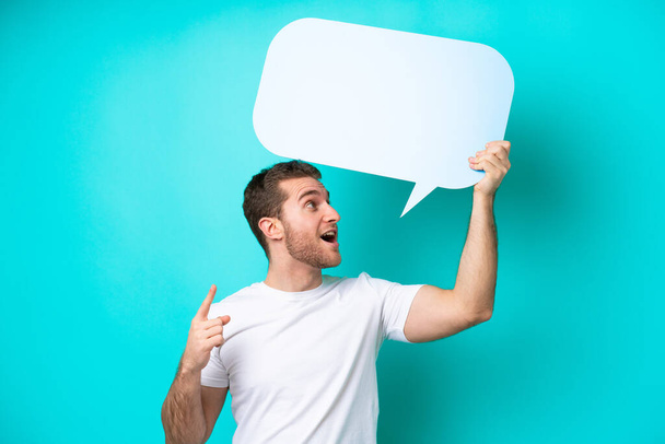 Young caucasian man isolated on blue background holding an empty speech bubble and thinking - Foto, imagen