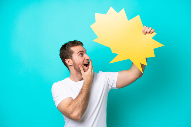 Young caucasian man isolated on blue background holding an empty speech bubble with surprised expression - Foto, imagen