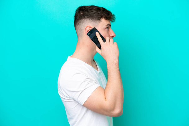 Jeune homme beau caucasien isolé sur fond bleu en gardant une conversation avec le téléphone mobile avec quelqu'un - Photo, image