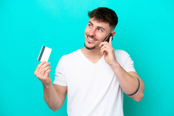 Jeune homme beau caucasien isolé sur fond bleu gardant une conversation avec le téléphone mobile et tenant une carte de crédit - Photo, image