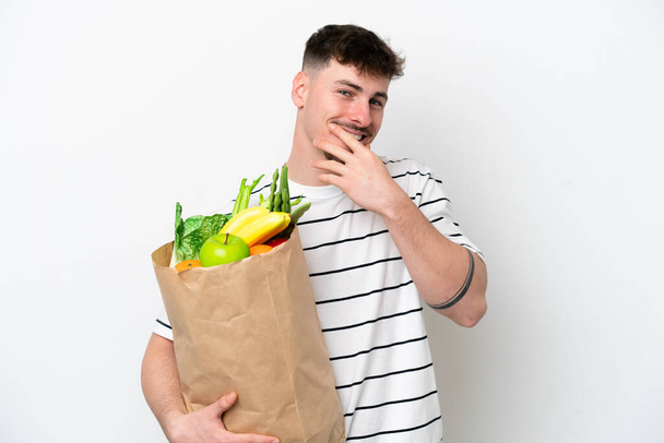 Joven hombre caucásico sosteniendo una bolsa de la compra de comestibles aislado sobre fondo blanco mirando hacia arriba mientras sonríe - Foto, Imagen