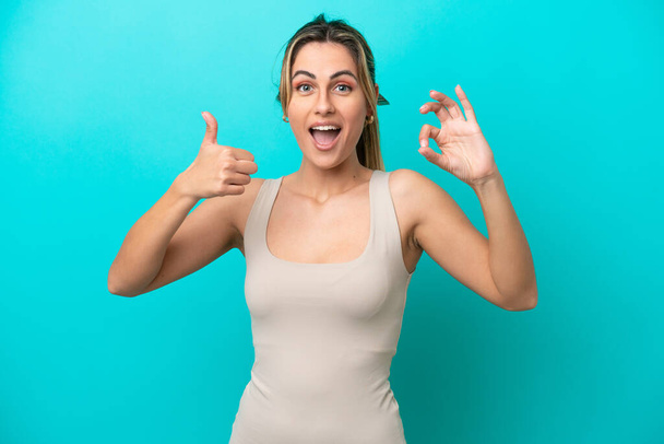 Young caucasian woman isolated on blue background showing ok sign and thumb up gesture - Fotoğraf, Görsel