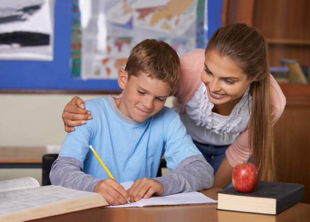 Cada niño es especial. Un joven profesor de apoyo se inclina sobre su estudiante mientras escribe en un libro. - Foto, Imagen