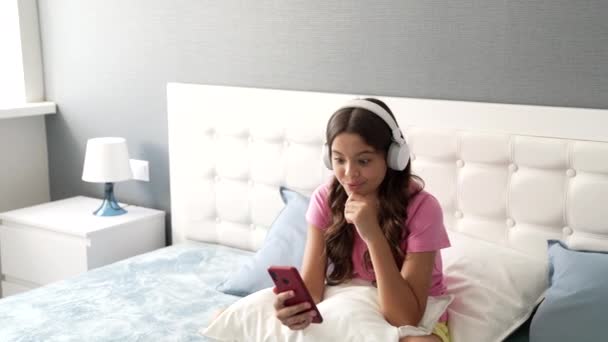niña feliz hablando en cámara de teléfono inteligente en auriculares para bloguear en el dormitorio, la vida moderna - Metraje, vídeo