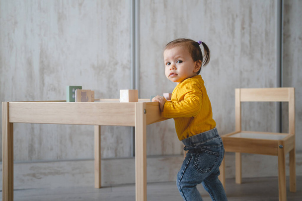 Ένα μωρό μικρό καυκάσιο κοριτσάκι παίζει στο τραπέζι στο σπίτι αντίγραφο χώρο κρατώντας ξύλινα παιχνίδια στο μπροστινό μέρος της ημέρας - Φωτογραφία, εικόνα
