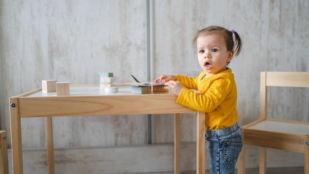 Um bebê pequena menina branca brincando na mesa no espaço de cópia em casa segurando brinquedos de madeira na vista lateral do dia - Foto, Imagem