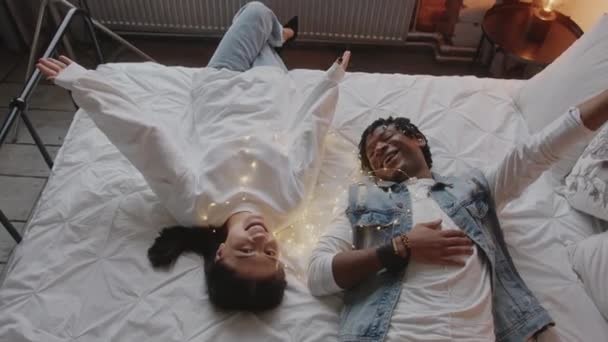 Couple mixte allongé sur leur lit king size, riant et profitant de leur temps libre. - Séquence, vidéo