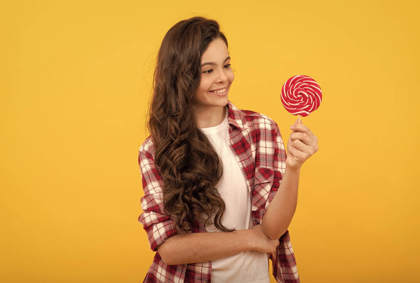 vrolijk kind met lang krullend haar houden lolly karamel snoep op gele achtergrond, tandheelkundige gezondheid. - Foto, afbeelding