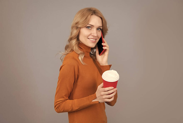 Happy girl talk on mobile phone holding hot cup of tea or coffee drink grey background, break - Photo, Image