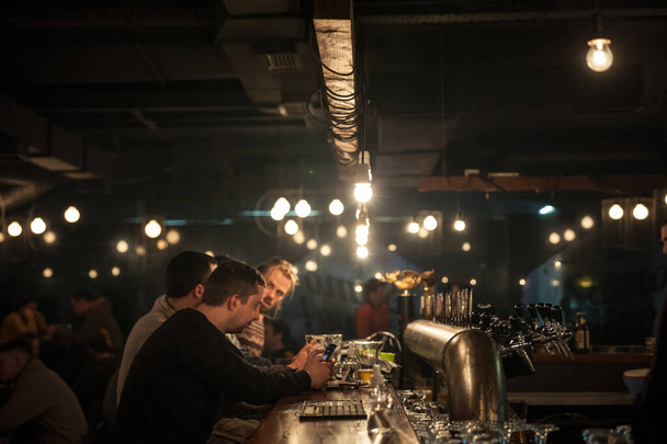 BELGRADE, SERBIA - DECEMBER 4, 2021: Selective blur on a man sitting at the counter of a pub, checking his smartphone, in front of tap beer, in a bar of belgrade. - Photo, Image