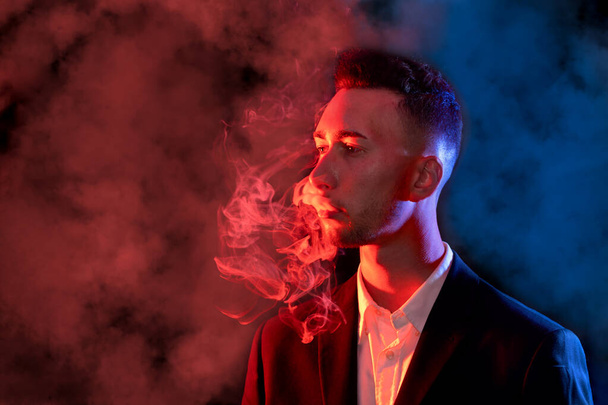 Elegantní mladý muž vyfukoval cigaretový kouř z úst na černém pozadí. Selektivní zaměření - Fotografie, Obrázek