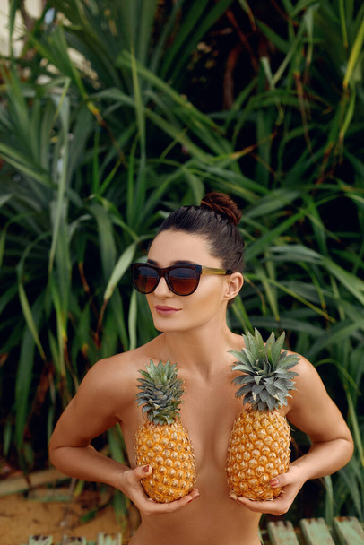 Young woman with pineapple in hand in the tropics. Beautiful nude girl with bronze tan in bikini holding a pineapple at the breast.  Covering her breast with pineapples - Foto, Imagen