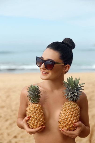 Beautiful caucasian sunbathed woman model with dark swimsuit posing on summer beach on blue sky and ocean background. Covering  her breast with pineapples - Photo, image