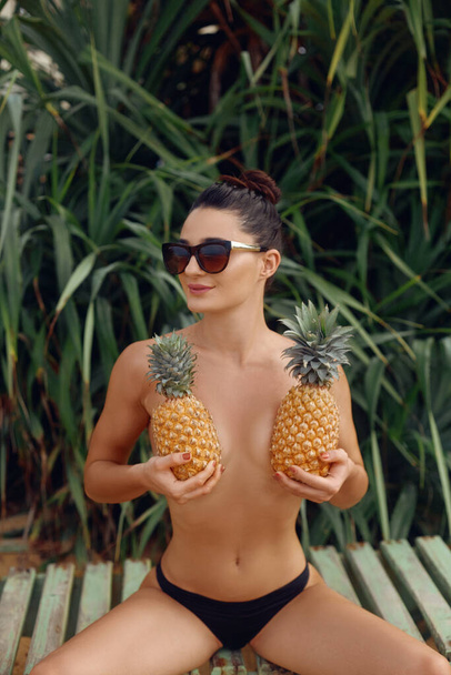 Young beautiful girl posing on the tropical background , holding pineapples in hands.  Woman with bronze tan in bikini. Covering her breast with pineapples - Photo, image