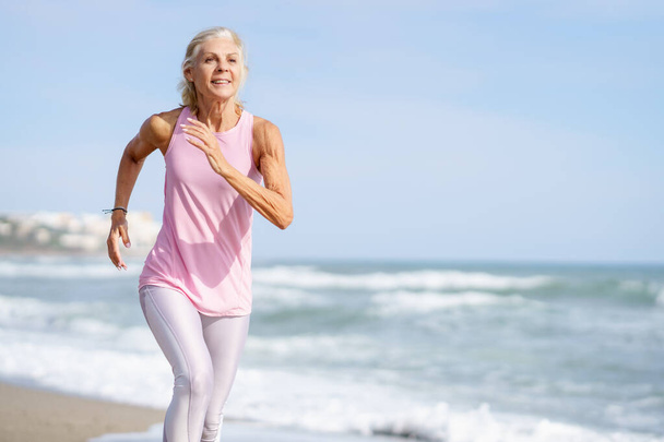 Felnőtt nő fut végig a part mentén. Idősebb nő sportol, hogy fitt maradjon. - Fotó, kép