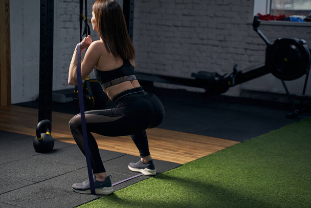 Athletic woman doing squats with fitness band in gym - Photo, Image