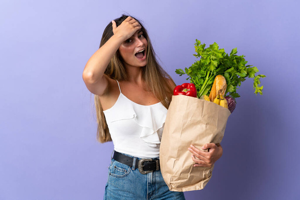 Mujer joven sosteniendo una bolsa de la compra de comestibles haciendo gesto sorpresa mientras mira a un lado
 - Foto, imagen
