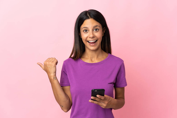 Valkoihoinen tyttö eristetty vaaleanpunainen tausta matkapuhelimella ja osoittaa sivusuunnassa - Valokuva, kuva
